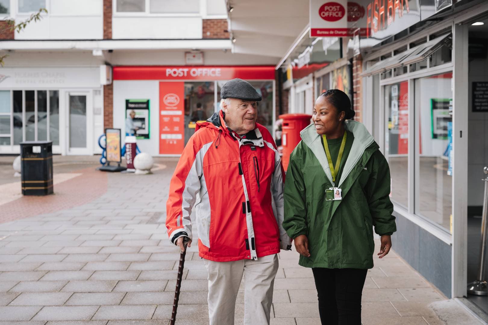 An elderly man with a walking stick walks outside with a carer. They are both smiling and talking. - Home Instead