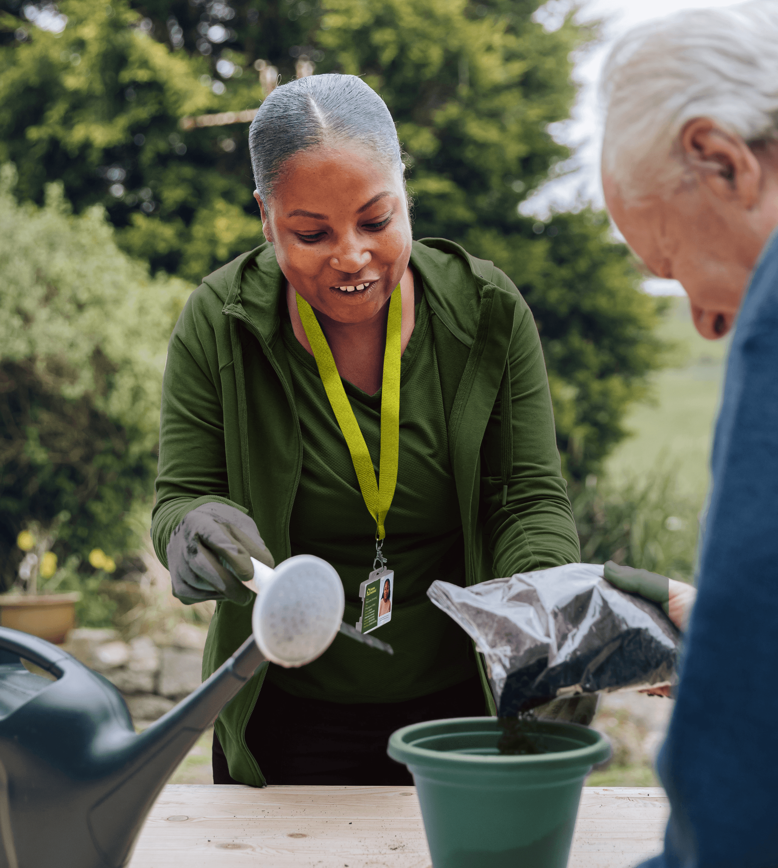 A woman helps an older man pot a plant outdoors, pouring compost into a green pot. - Home Instead