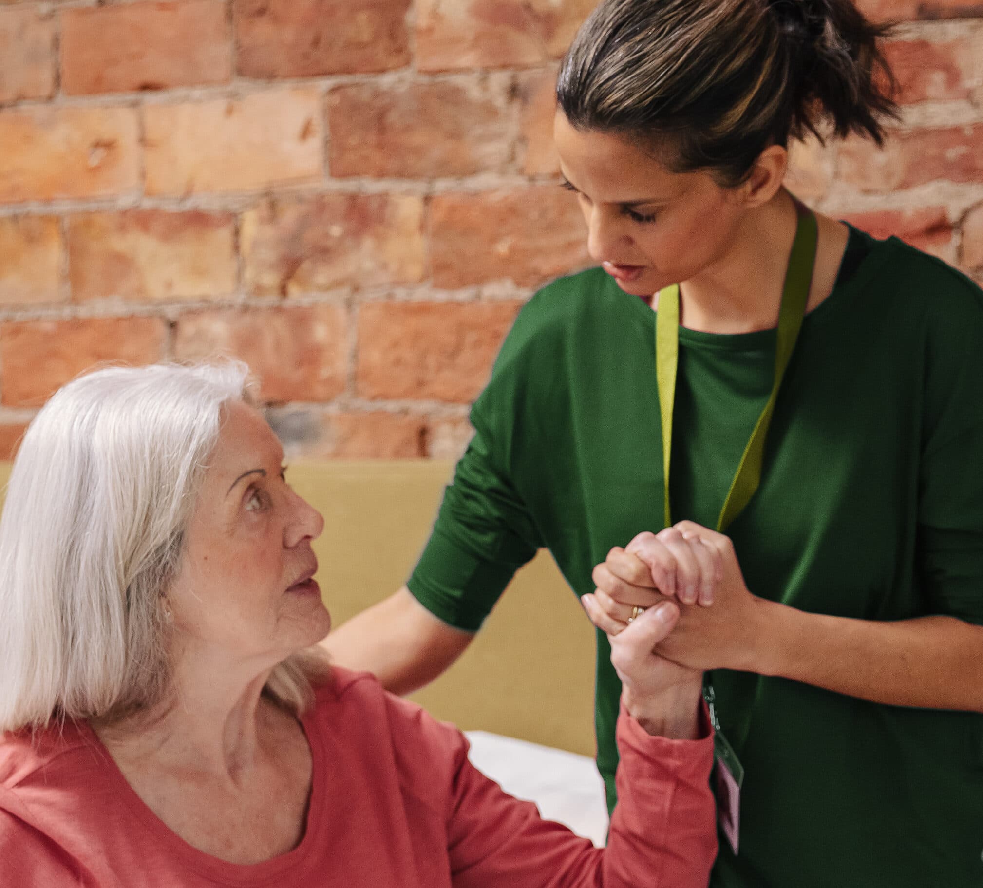 A caregiver in green supports an elderly woman in pink, holding her hand and making eye contact. - Home Instead