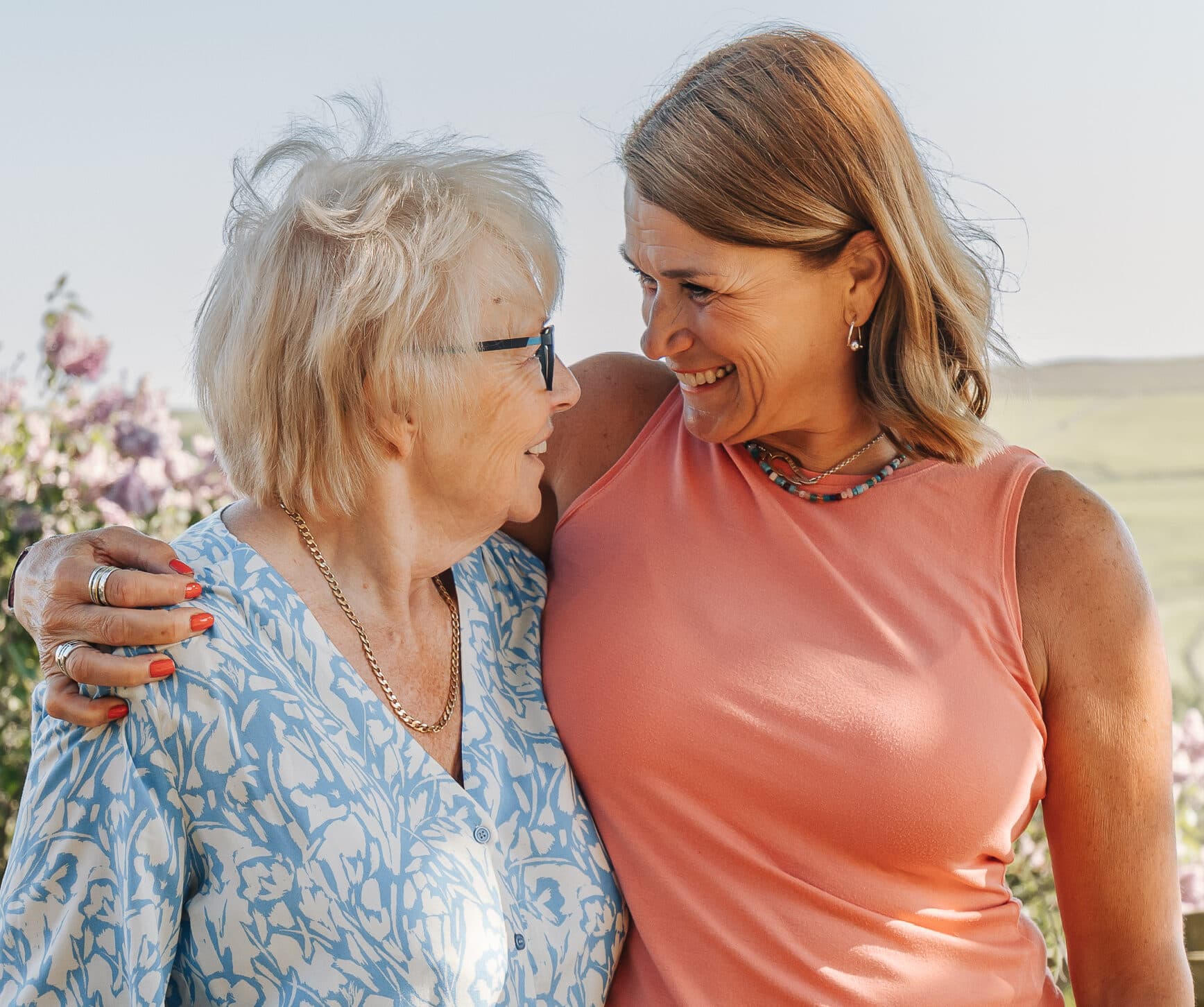 Two women smiling at each other outdoors, one older and one younger, with arms around each other. - Home Instead