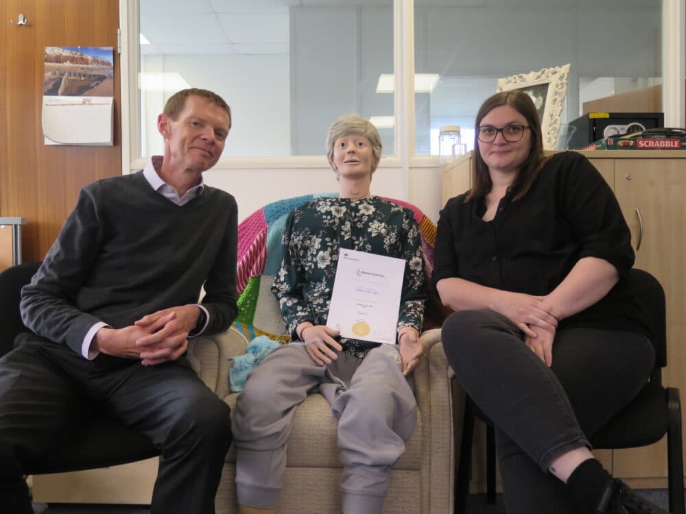 Two people sit beside a human-like mannequin holding a certificate, all smiling at the camera in an office setting. - Home Instead