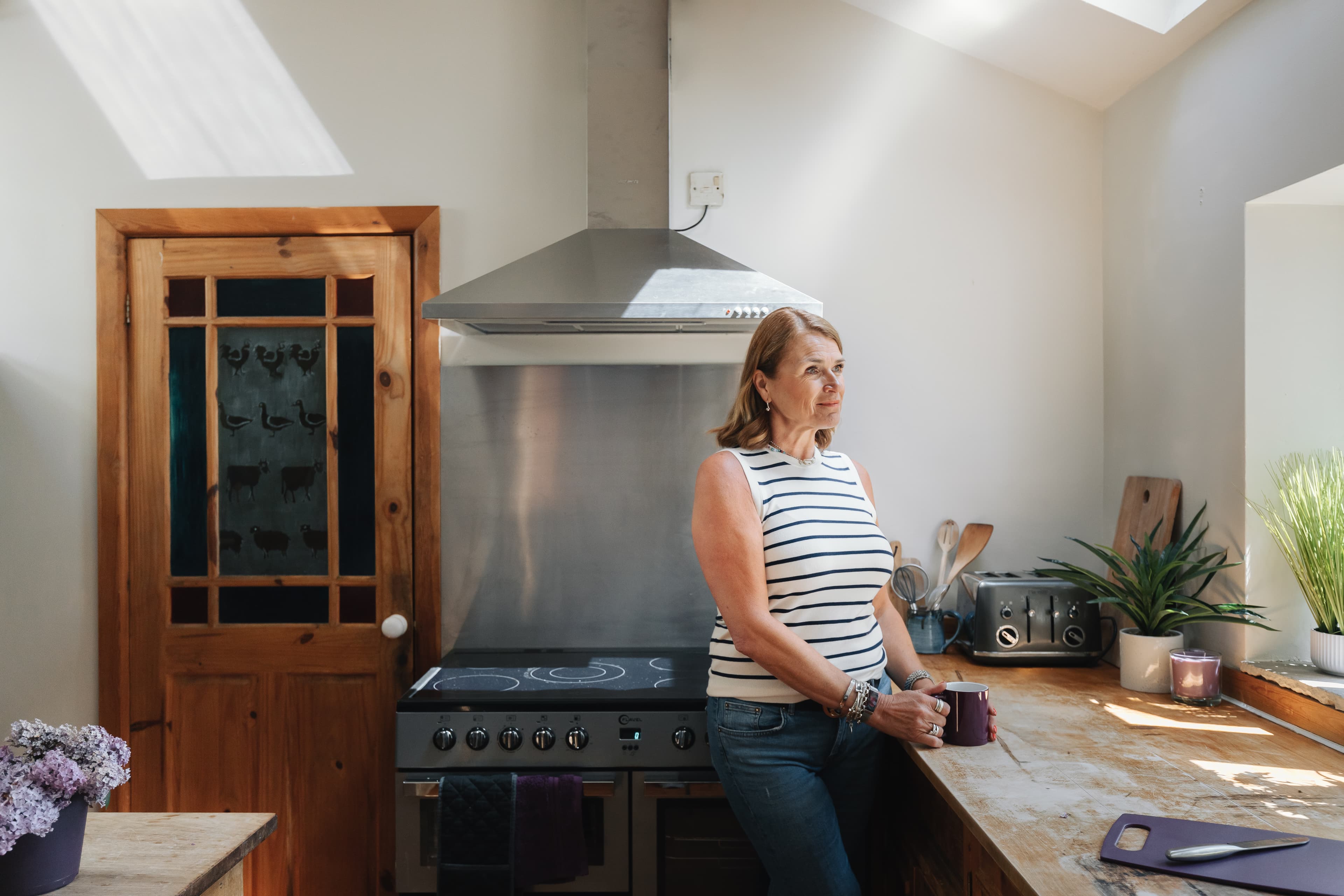 Woman in a striped top stands in a sunlit kitchen, holding a mug and looking thoughtfully out the window. - Home Instead