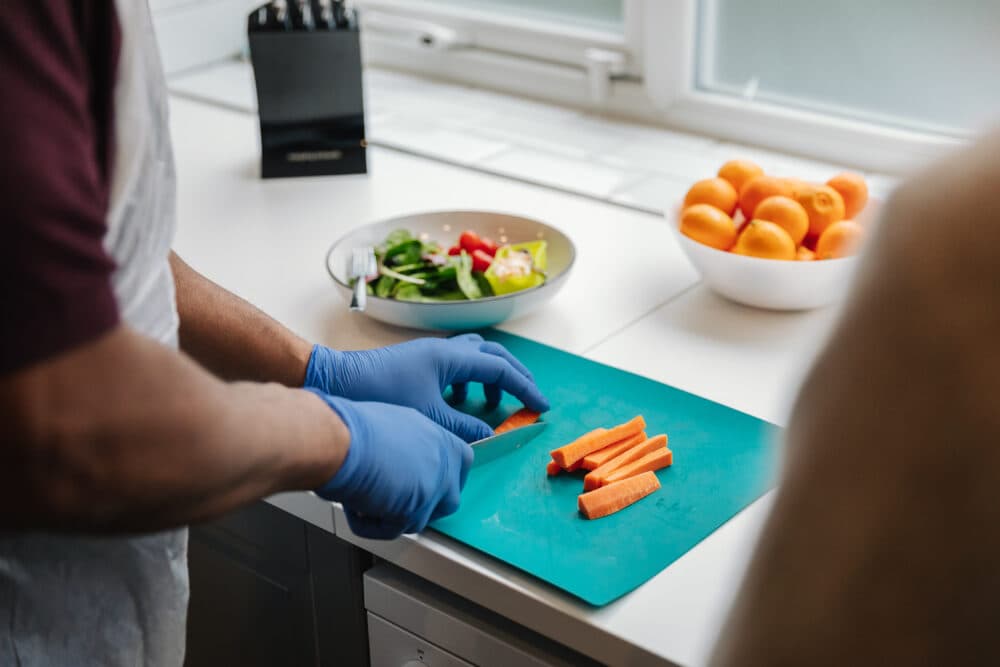 Person wearing blue gloves slicing carrots on a cutting board, with salad and fruit on the counter nearby. - Home Instead