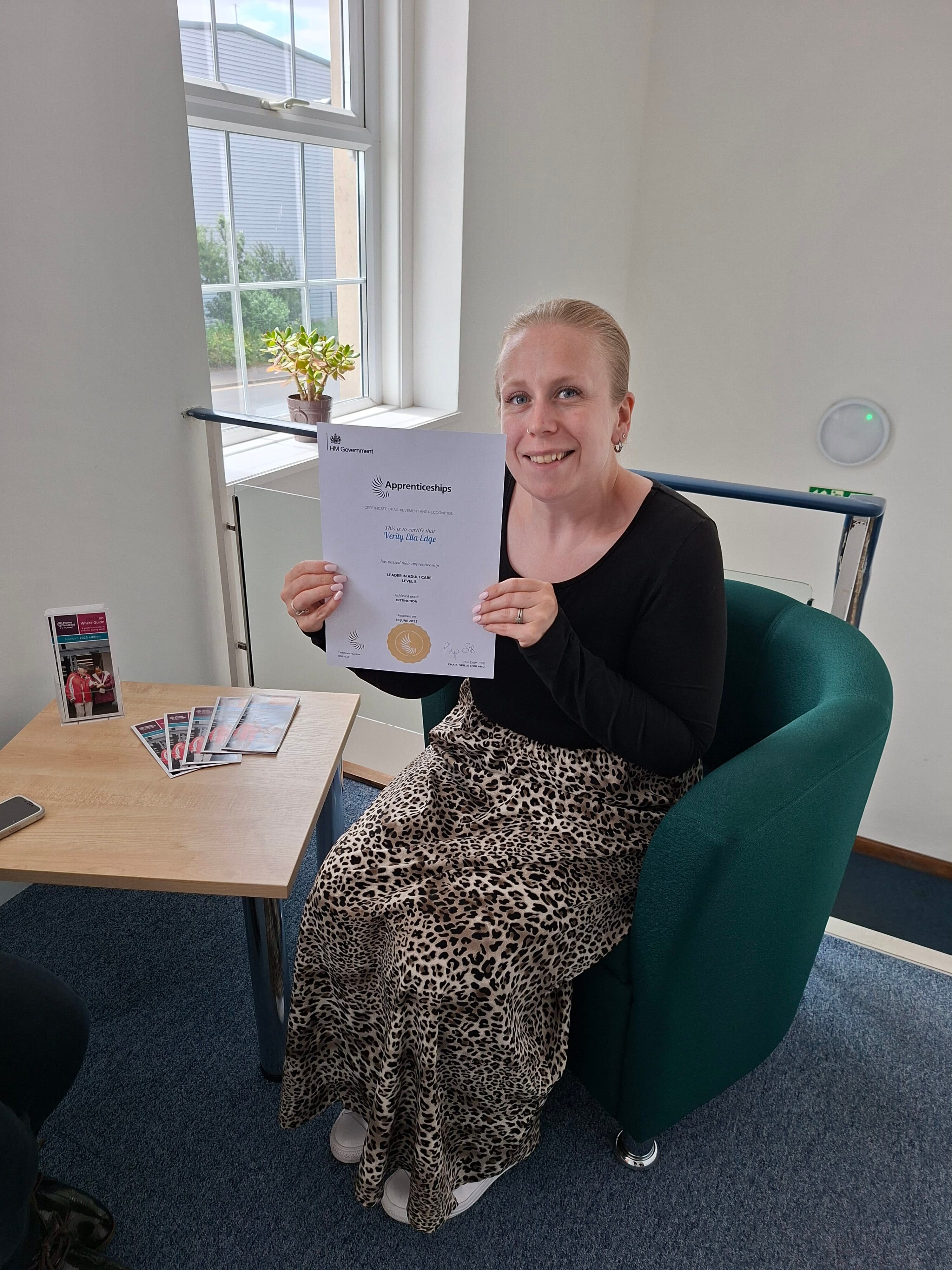 Woman sitting and smiling, holding an apprenticeship certificate, next to a table with brochures. - Home Instead