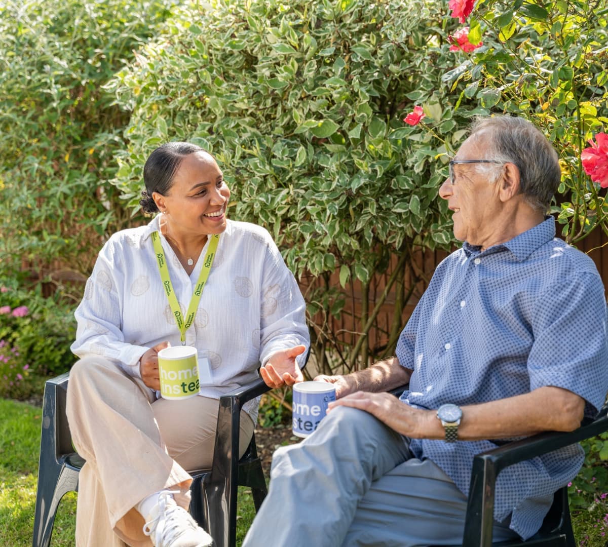 A woman and an elderly man sit outside, smiling and talking, each holding a mug in a garden setting. - Home Instead