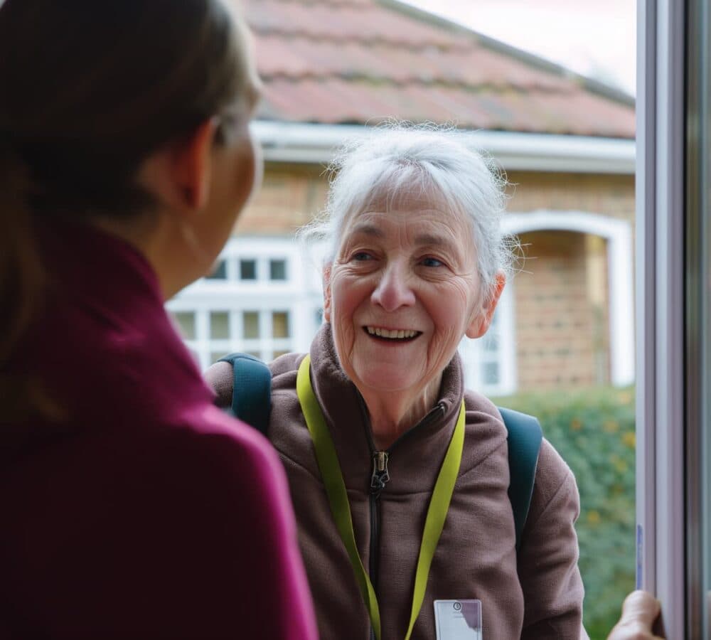 An older woman smiling and talking to another person at a door, wearing a lanyard and brown jacket. - Home Instead