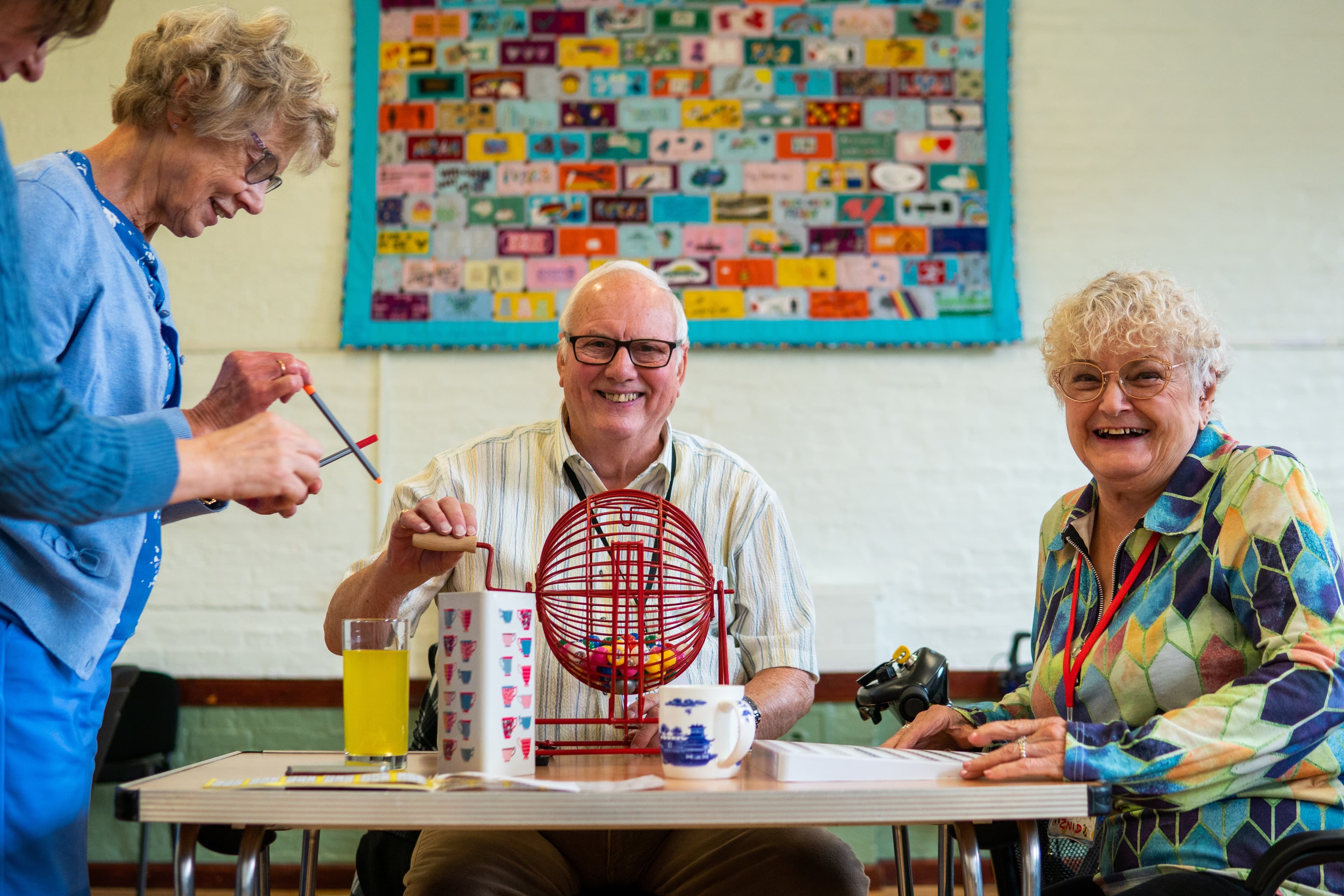 Three older adults sitting together at a table, smiling and playing a game of bingo. One man is turning a red bingo cage while two women sit nearby, one with a colorful patterned blouse and glasses, and another standing with pens in hand. Drinks and mugs are on the table, and a colorful patchwork quilt hangs on the wall behind them.