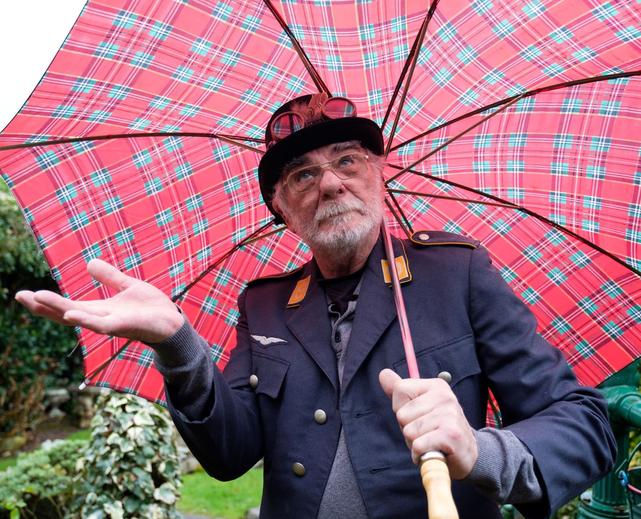Elderly man with a tartan umbrella and wearing a uniform, brimmed hat and goggles on the hat, wondering whether it is raining.
