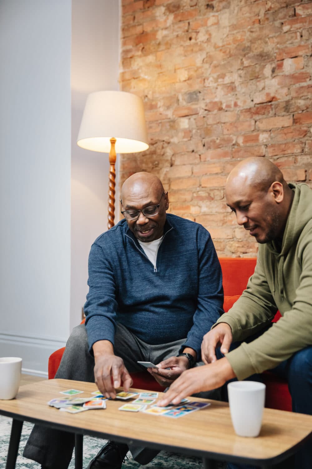 Two men sit on a red couch playing cards at a coffee table with two white mugs in a cozy room. - Home Instead