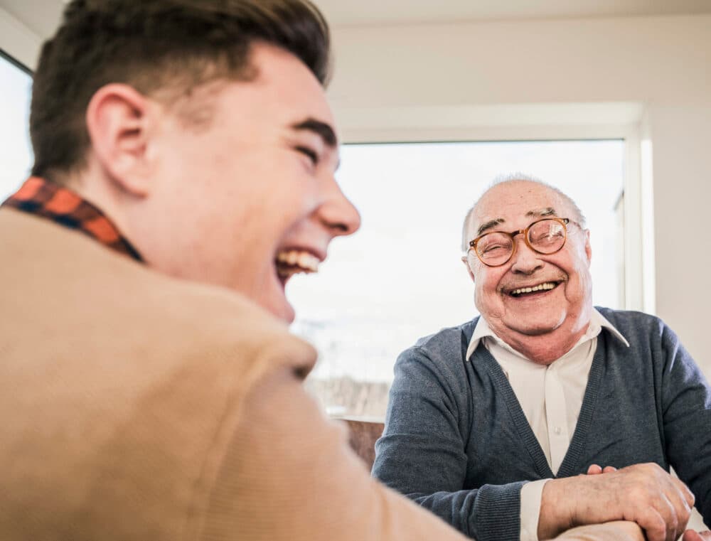 Young man and elderly man laughing together at a table in a bright room. - Home Instead