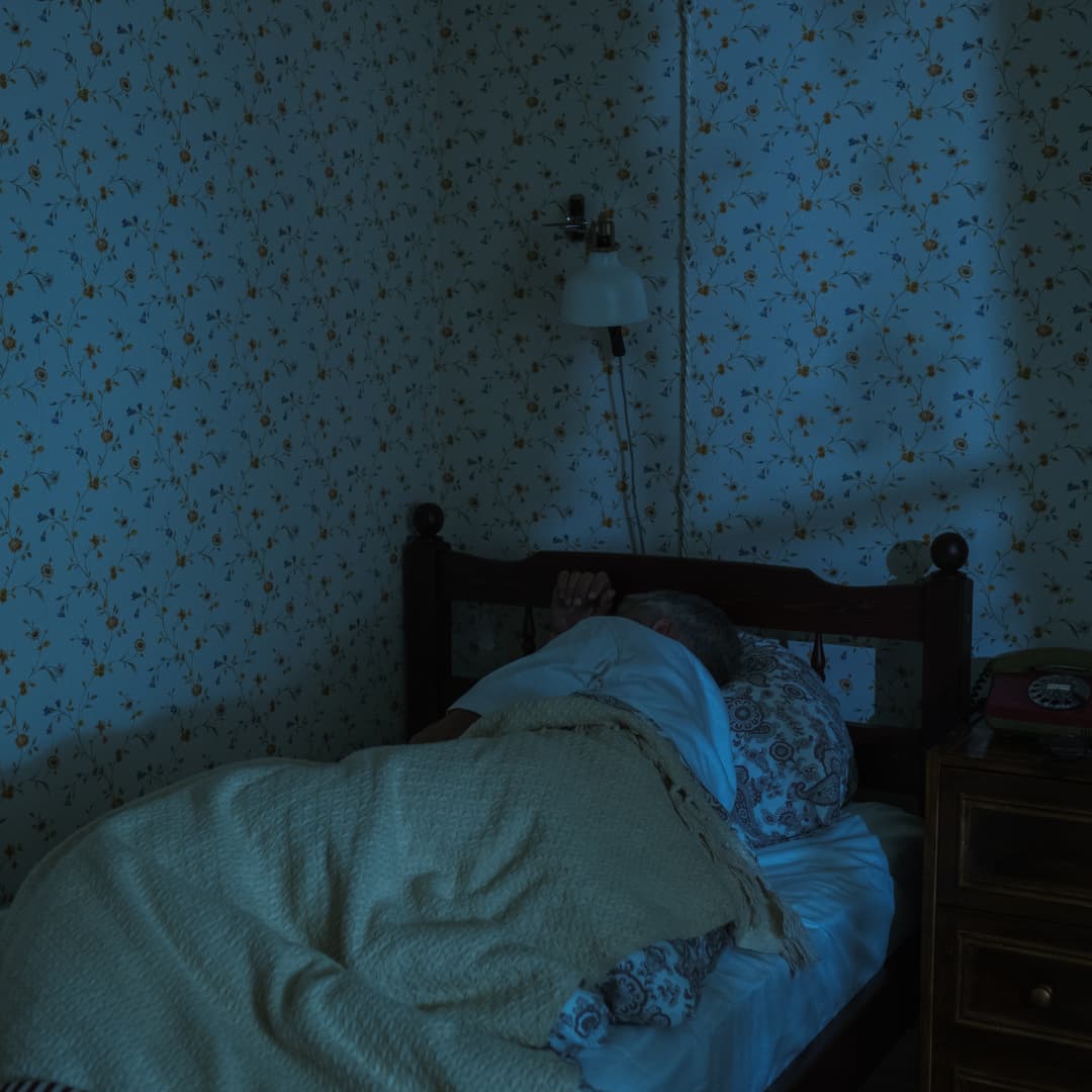 A person lies in bed under a blanket in a dimly lit room with floral wallpaper. - Home Instead