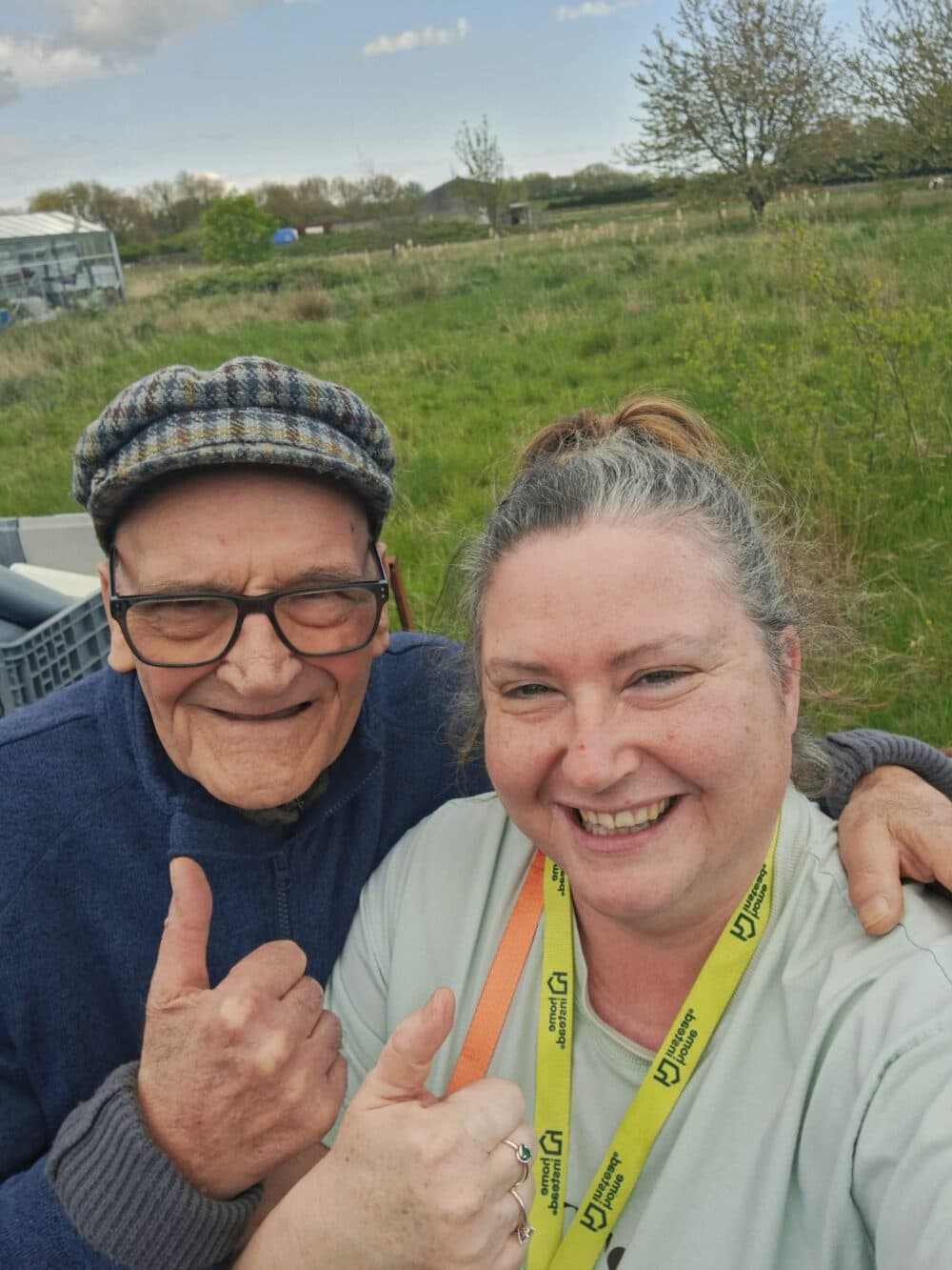 An elderly man and a woman outdoors smiling and giving thumbs up, with green fields in the background. - Home Instead