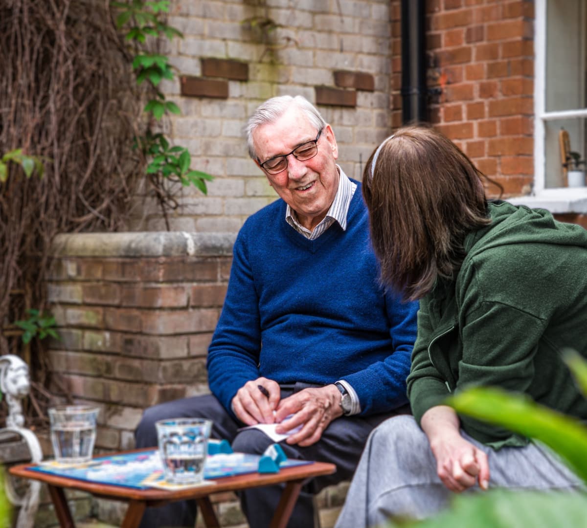 An older male adult wearing eyeglasses and blue sweater happy and smiling while chatting with his female carer outside the house