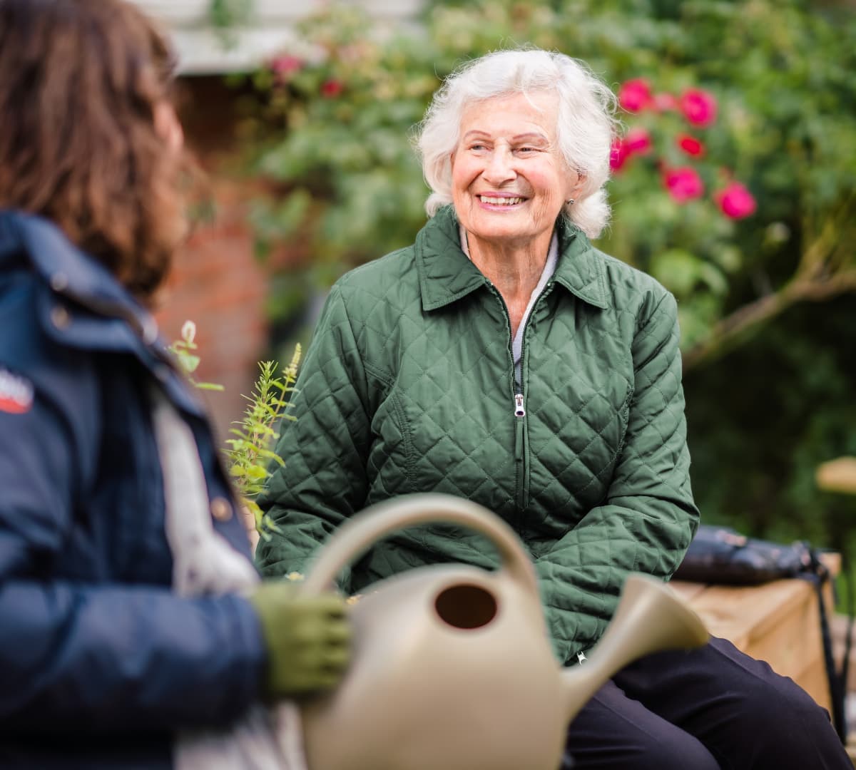 Smiling elderly woman in a green jacket sits outdoors, talking to another person holding a watering can. - Home Instead