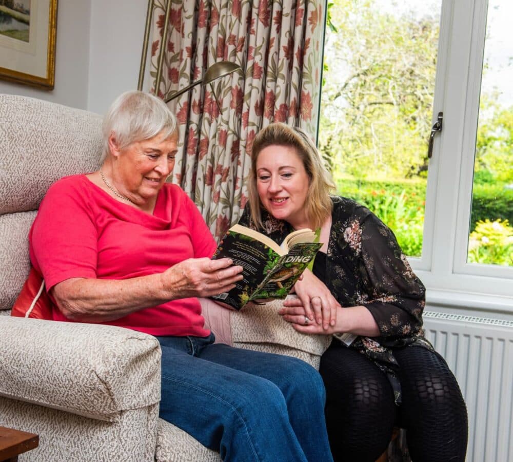 An older female adult sitting on a couch while reading a book by the window with her younger female carer both happy and smiling