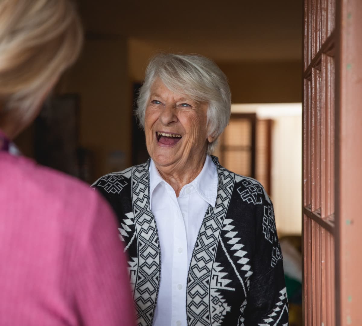 An older female adult with grey hair and wearing a sweater happy and smiling while at the door and greeting someone - Home Instead