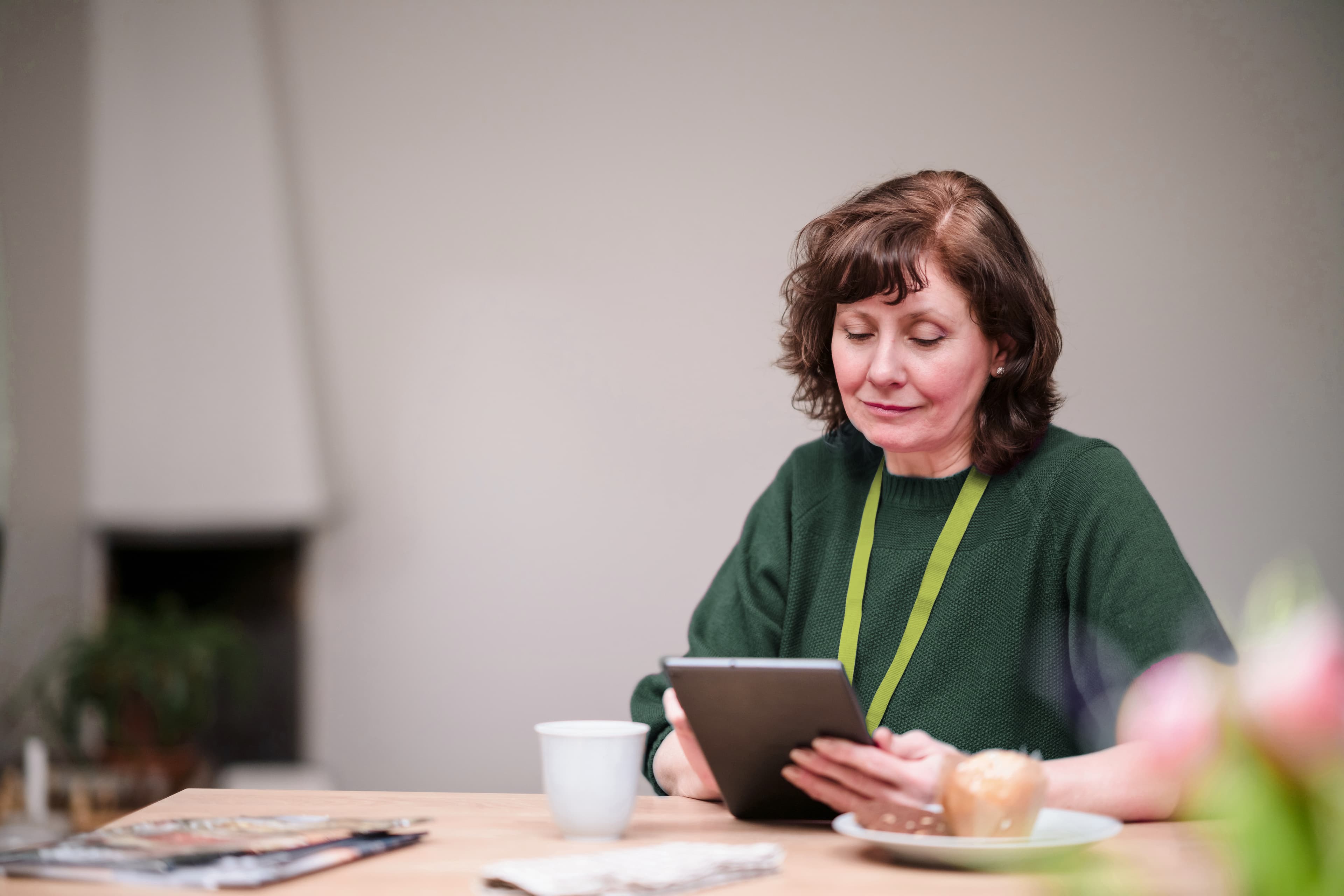 Woman with brown hair in a green sweater reads a tablet at a table with coffee and magazines. - Home Instead