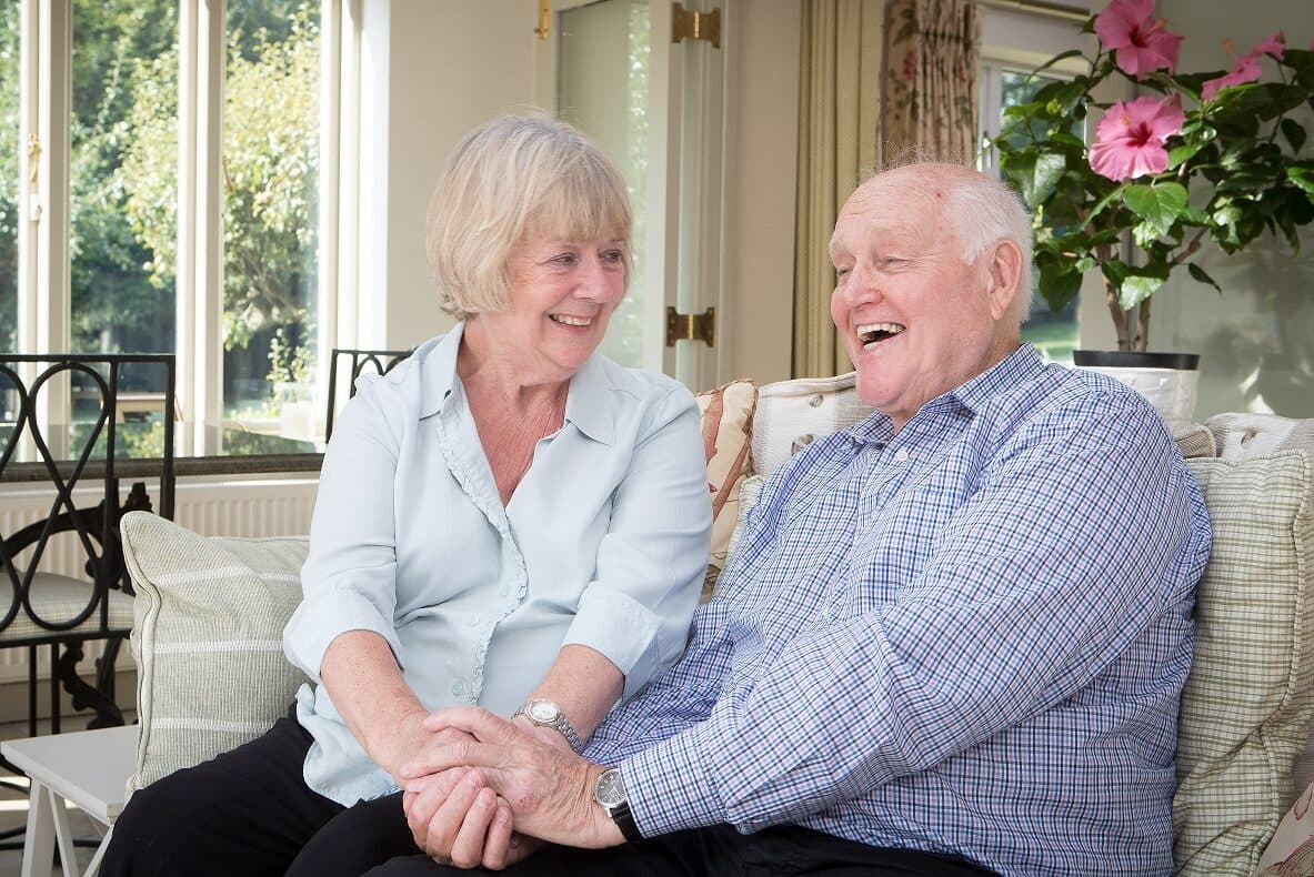 Elderly couple sitting on a sofa holding hands and looking at each other, laughing