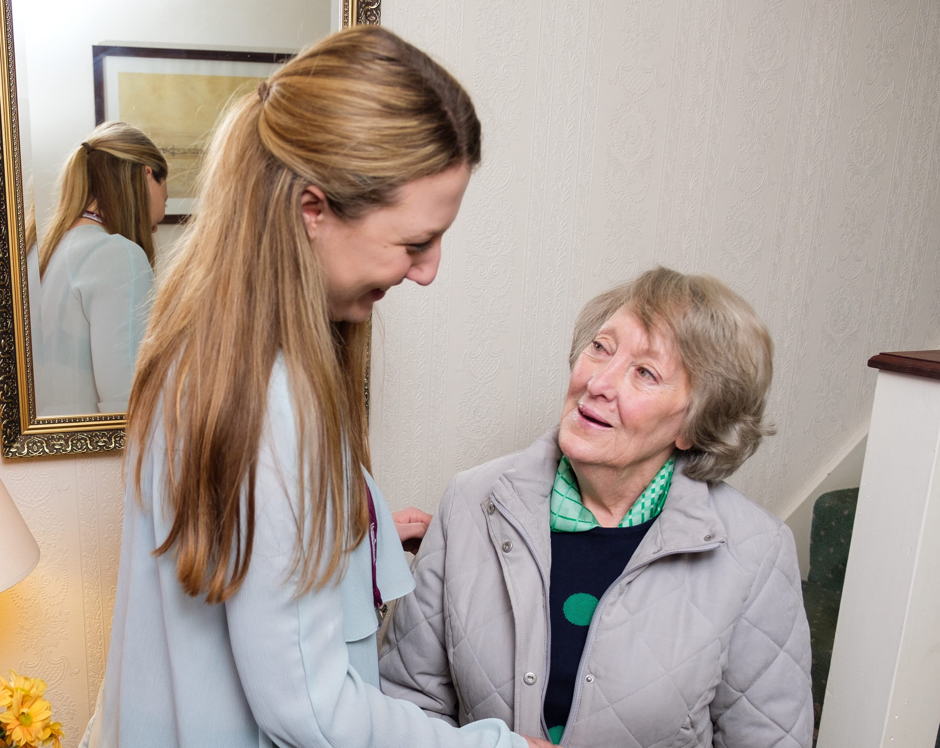 Young female carer helping her elderly female client to put on her coat, standing in the hallway of the client's home.