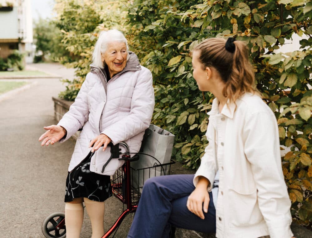 Older woman sitting on a walker, talking and smiling with a younger woman outdoors near green bushes. - Home Instead