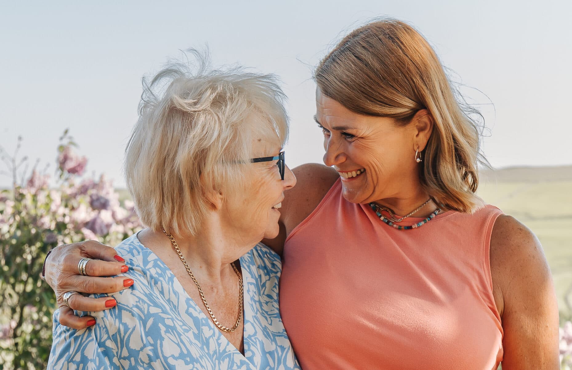 Two women smiling and embracing outdoors, looking at each other warmly on a sunny day. - Home Instead