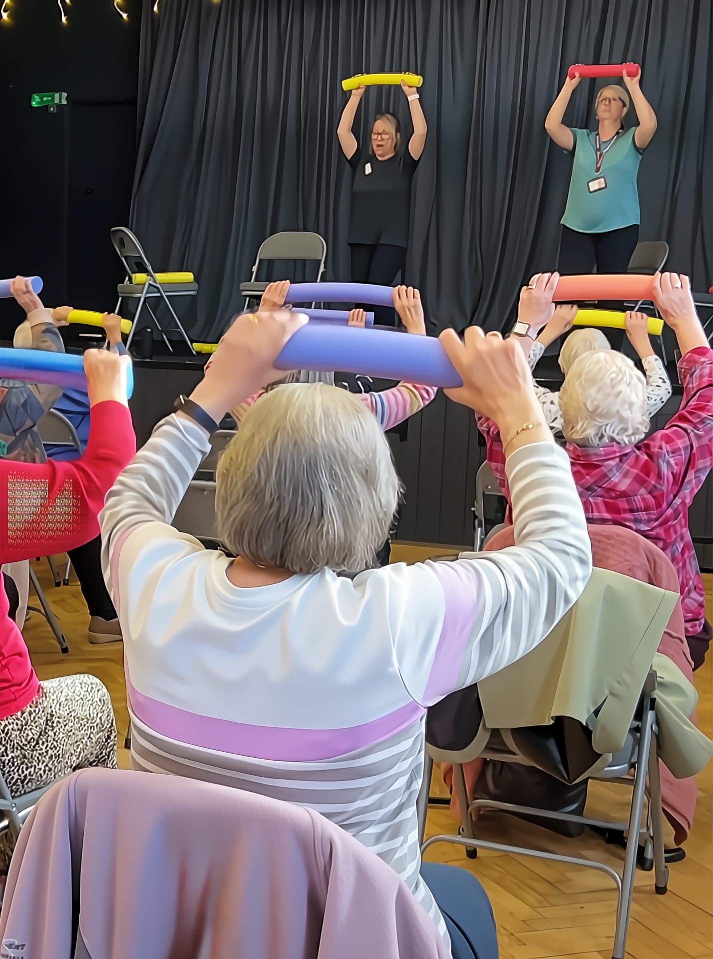Women at a seated exercise class exercising with baton and following two female instructors on the stage