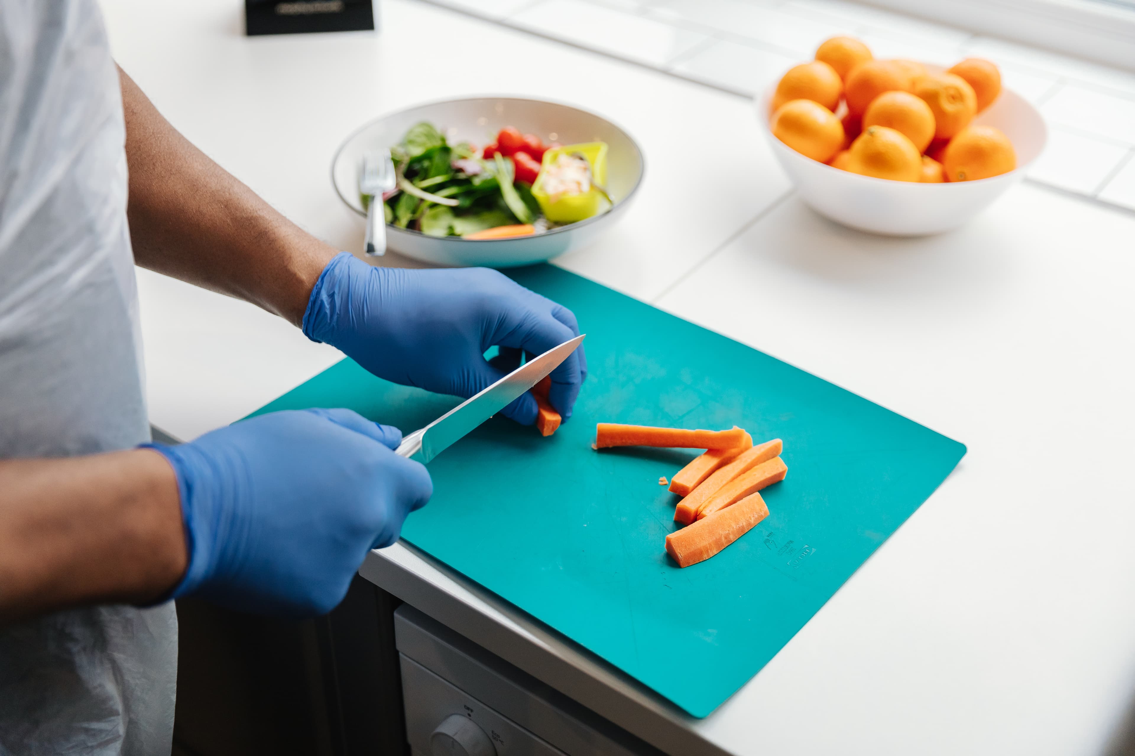 Person wearing blue gloves slicing carrots on a teal chopping board beside a bowl of salad and oranges. - Home Instead