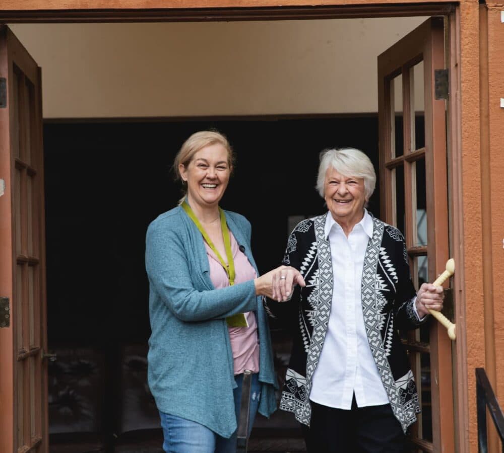 An older female adult going out of the door while being helped by a younger female carer both happy and smiling