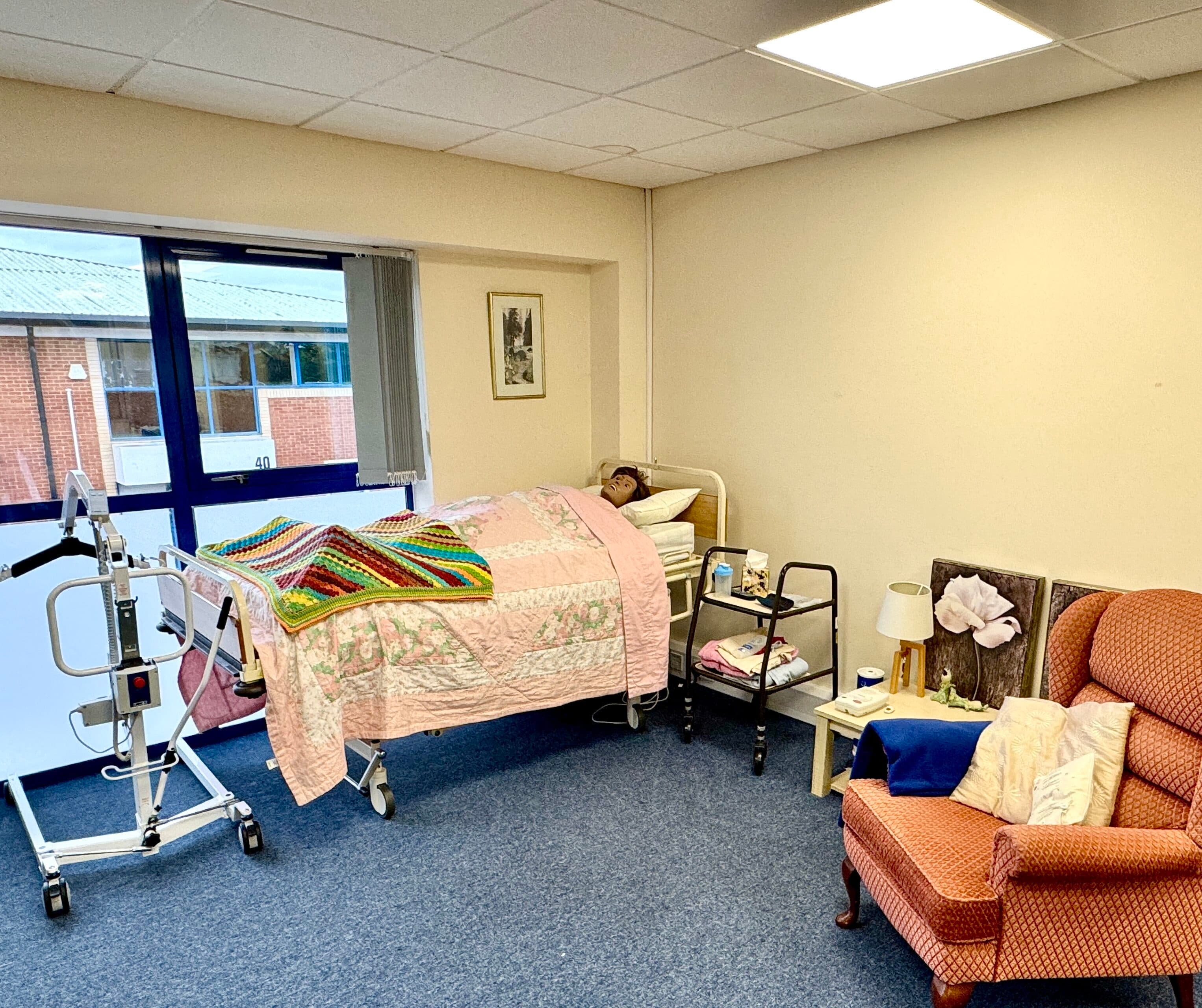 A person lies in a hospital bed in a bright room with medical equipment, a chair, and bedside tables. - Home Instead