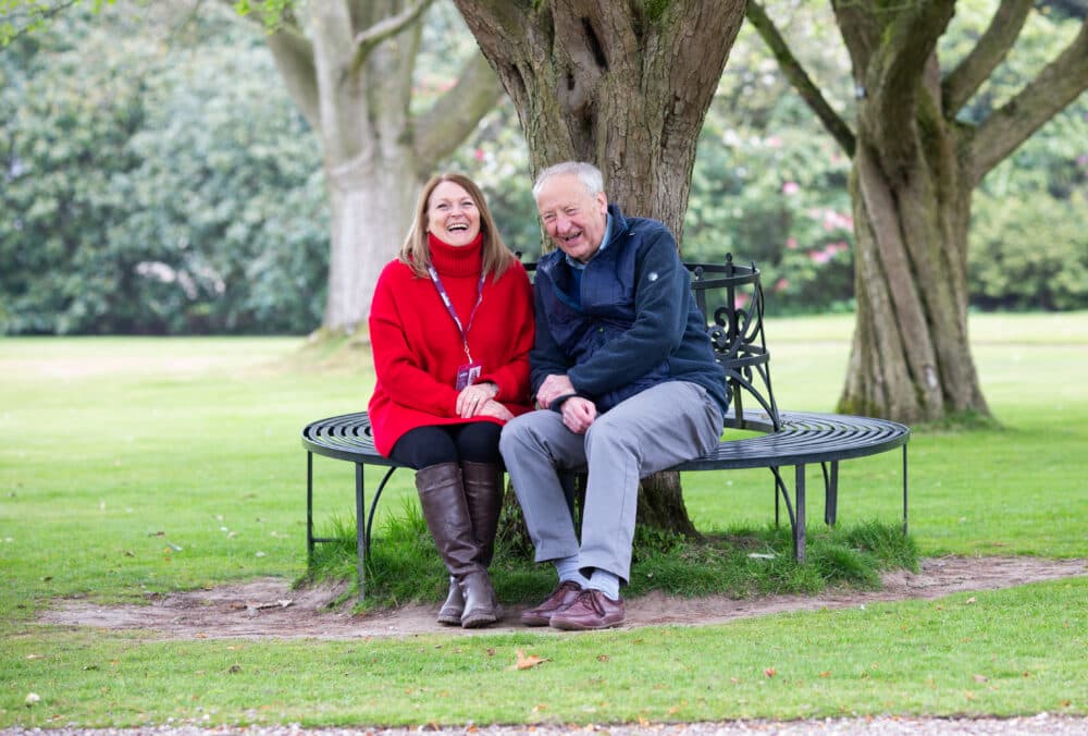 Elderly man and his female carer sitting on a circular bench around a large tree in a park, looking happy