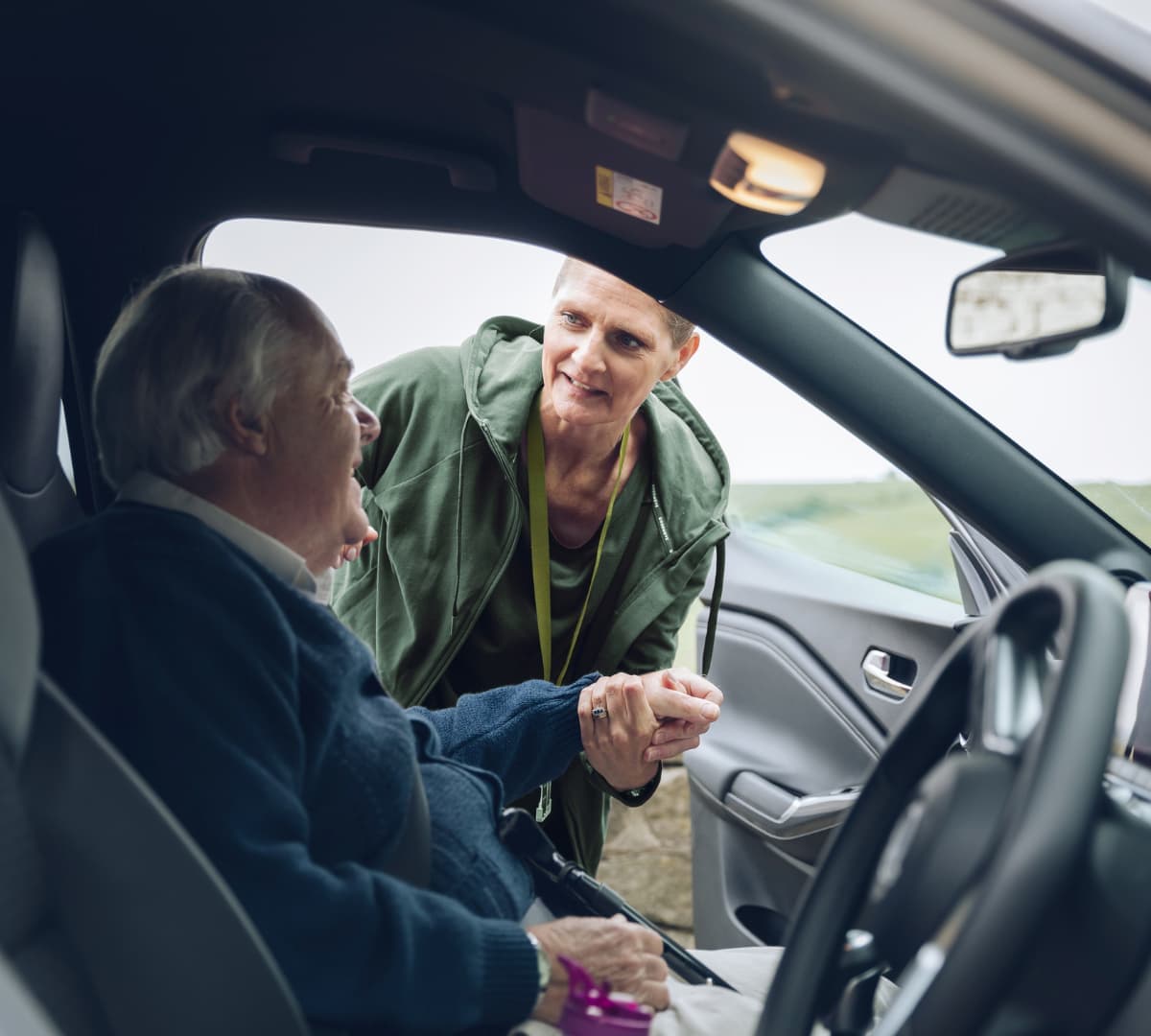 An older male adult with grey hair and wearing blue sweater going out of the car while being helped by a younger female carer wearing green