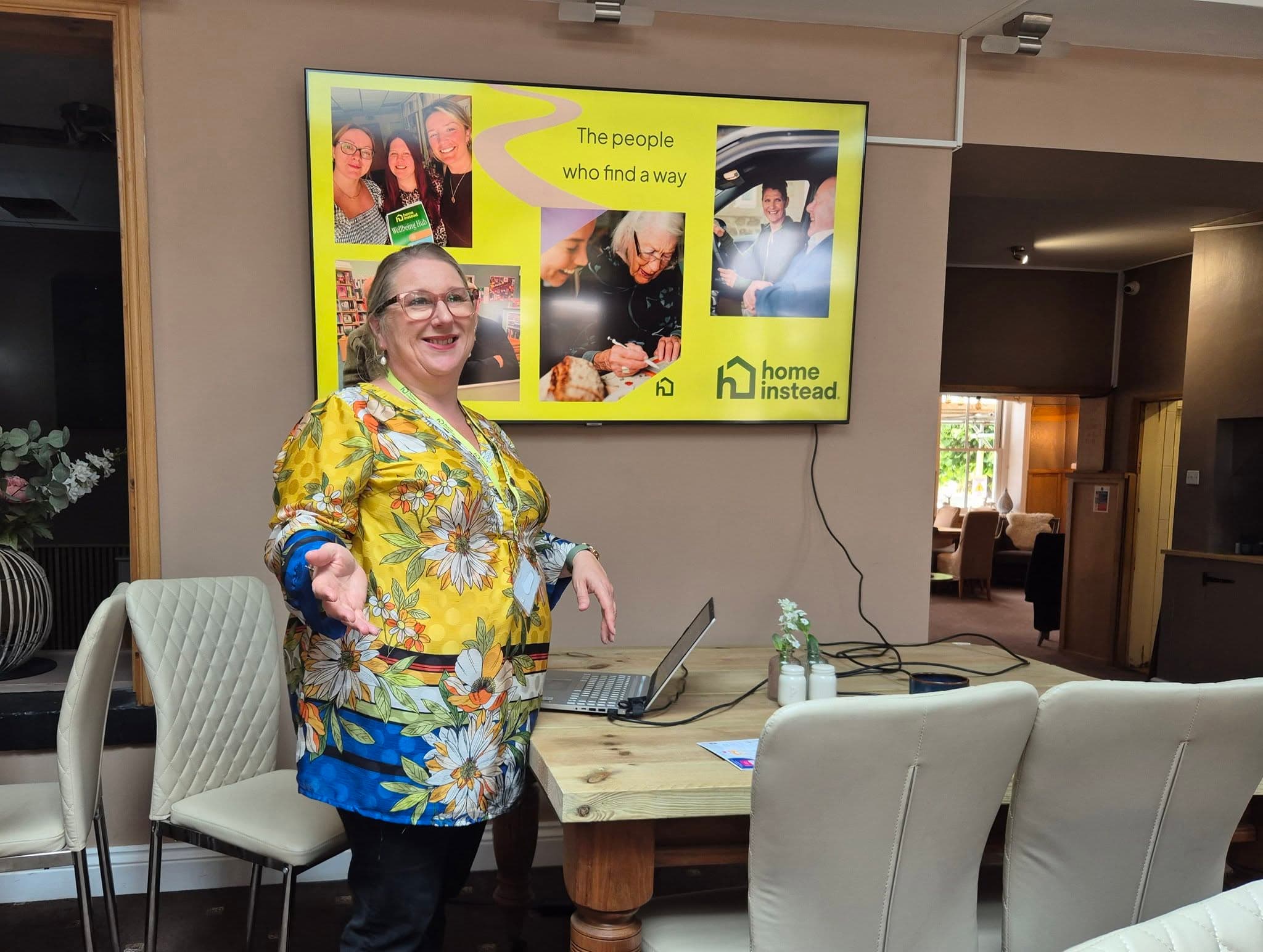A woman in a floral shirt presents by a screen with "The people who find a way" and "home instead" displayed. - Home Instead