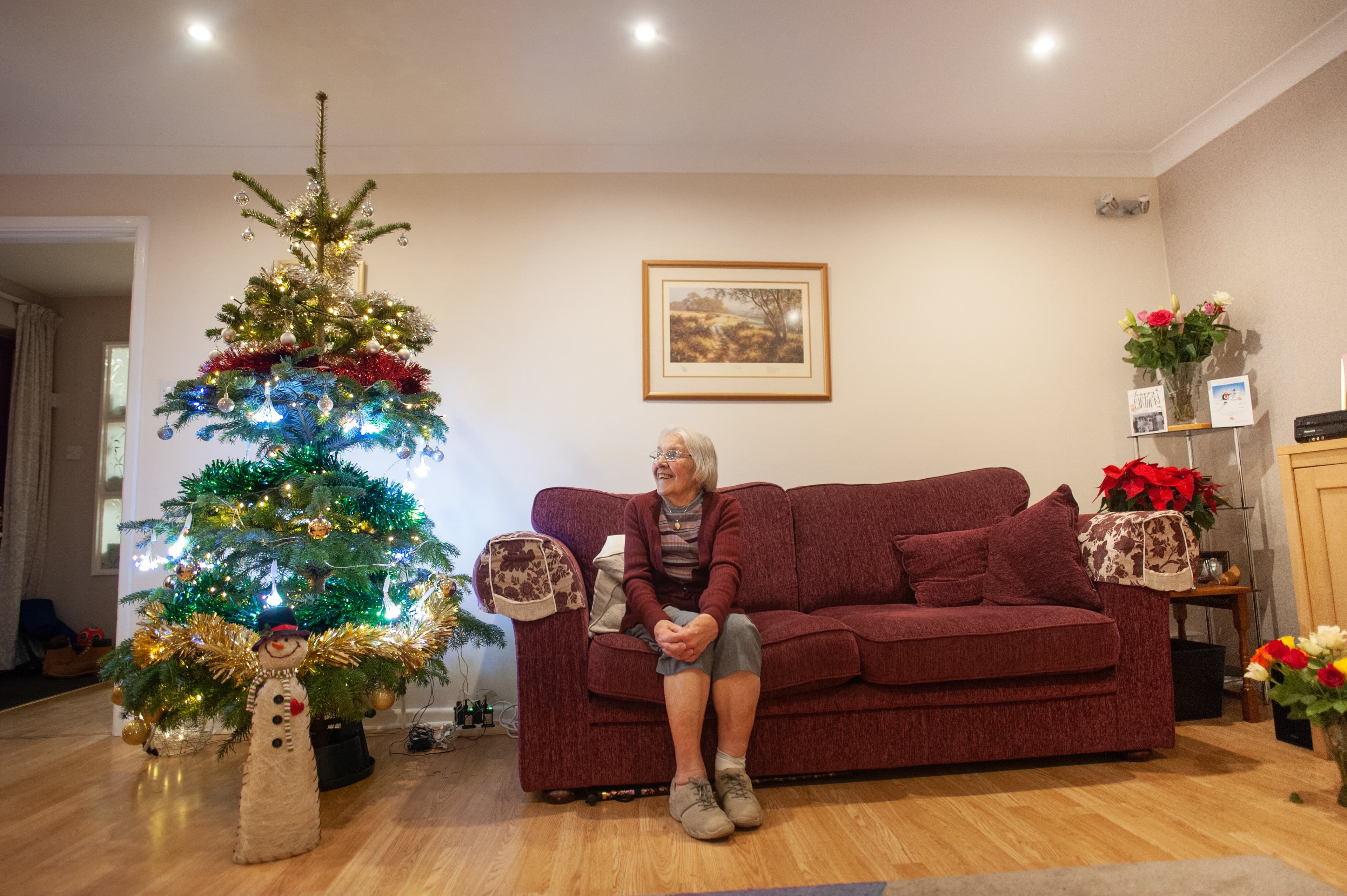 An older woman sits on a couch by a decorated Christmas tree in a cozy living room. - Home Instead