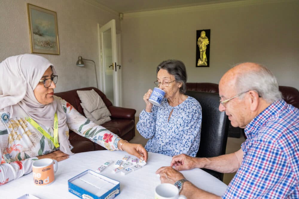 Three seniors sit at a table playing cards, drinking from mugs, and enjoying each other's company in a cozy room. - Home Instead