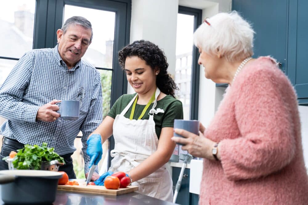 A young woman chops vegetables in a kitchen with two older adults, all smiling and holding mugs. - Home Instead