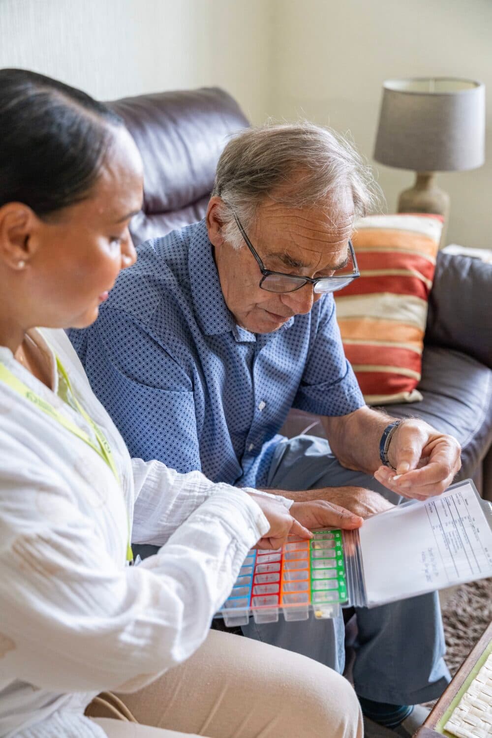 An elderly man reviews a medication schedule with a caregiver, using a colorful pill organizer. - Home Instead