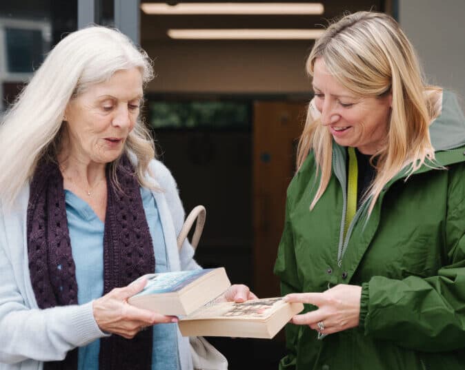 Two women standing outside, smiling and exchanging books with each other. - Home Instead