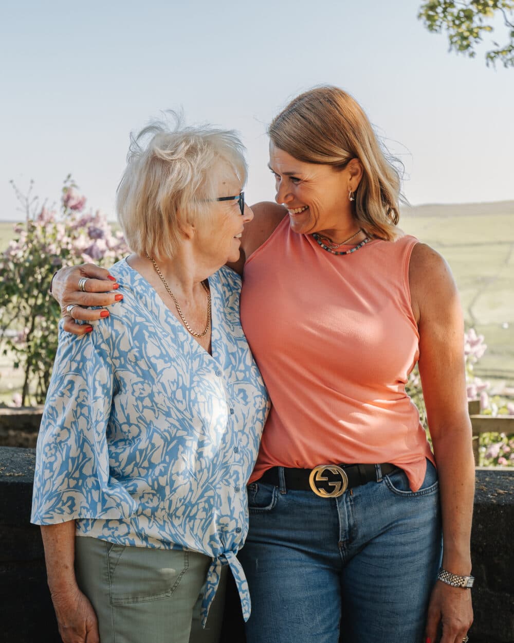 Two women standing outdoors smiling at each other, one with her arm around the other. - Home Instead