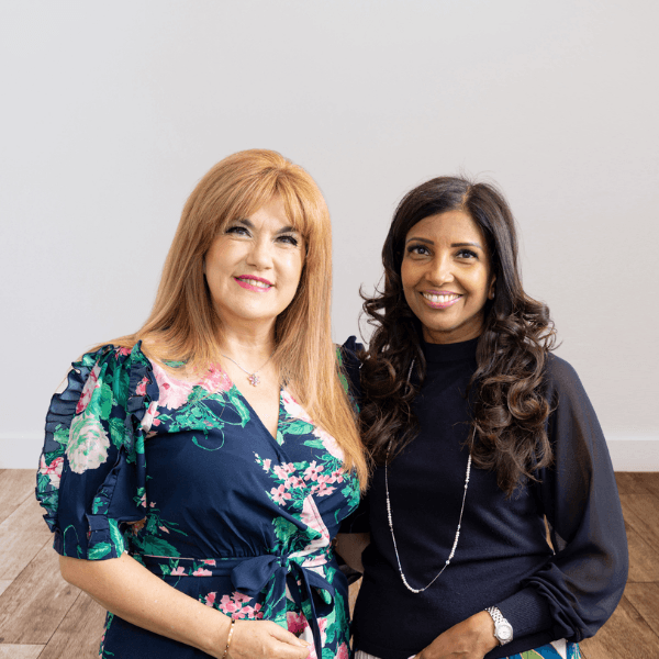 Two women standing together and smiling in a bright room with a wooden floor and light-colored walls. - Home Instead