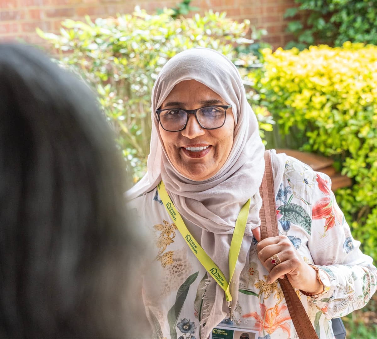 A female carer wearing turban and eyeglasses and floral dress smiling at the door while carrying a bag