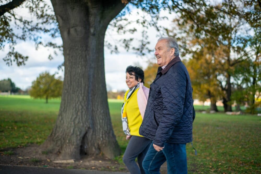 Two older adults walking together in a park on a cloudy day, smiling near a large tree. - Home Instead