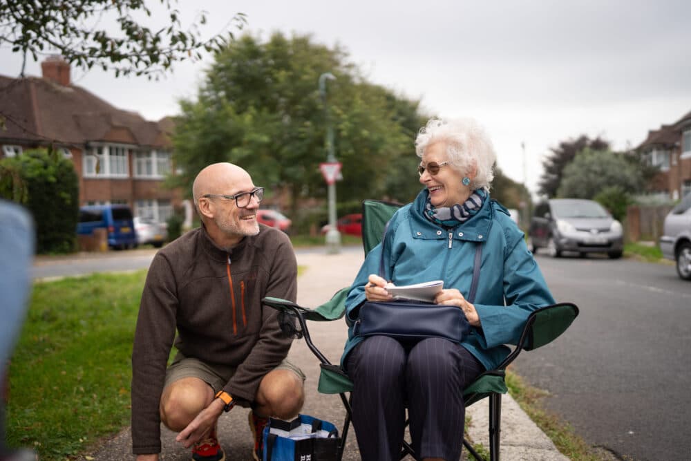 An older woman sits in a chair outdoors, smiling and talking with a man kneeling beside her on a suburban street. - Home Instead