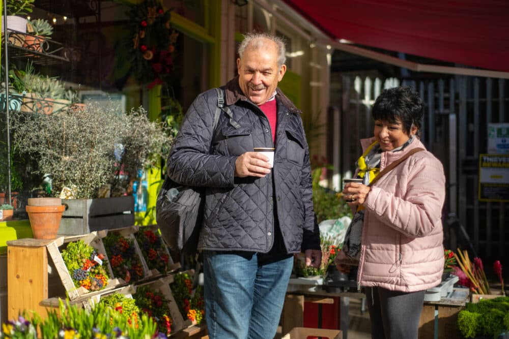 Two people smiling and holding drinks, standing outside a flower shop on a sunny day. - Home Instead