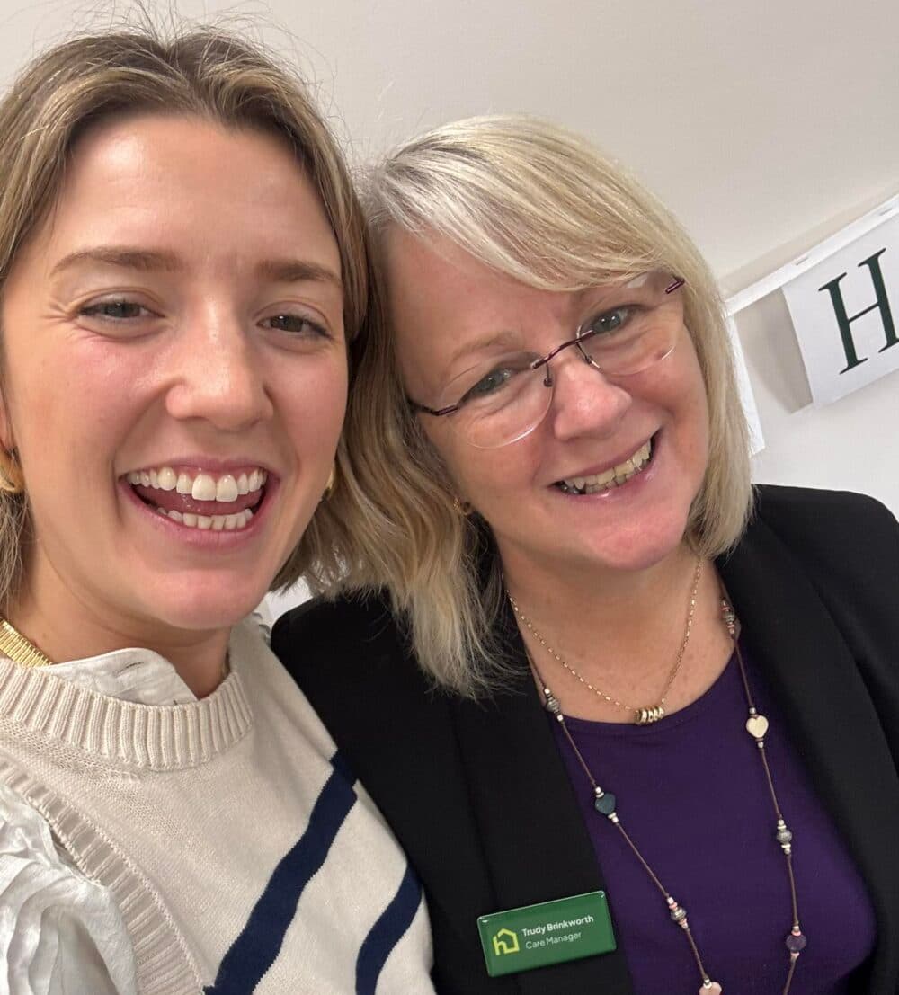 Two women smiling together indoors; one wears a cream jumper, the other a name badge and glasses. - Home Instead