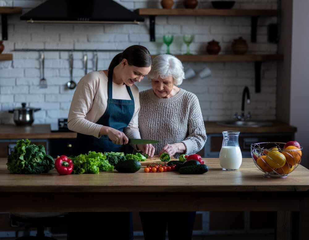 A young woman and an elderly woman prepare vegetables together in a kitchen, smiling and enjoying the moment. - Home Instead