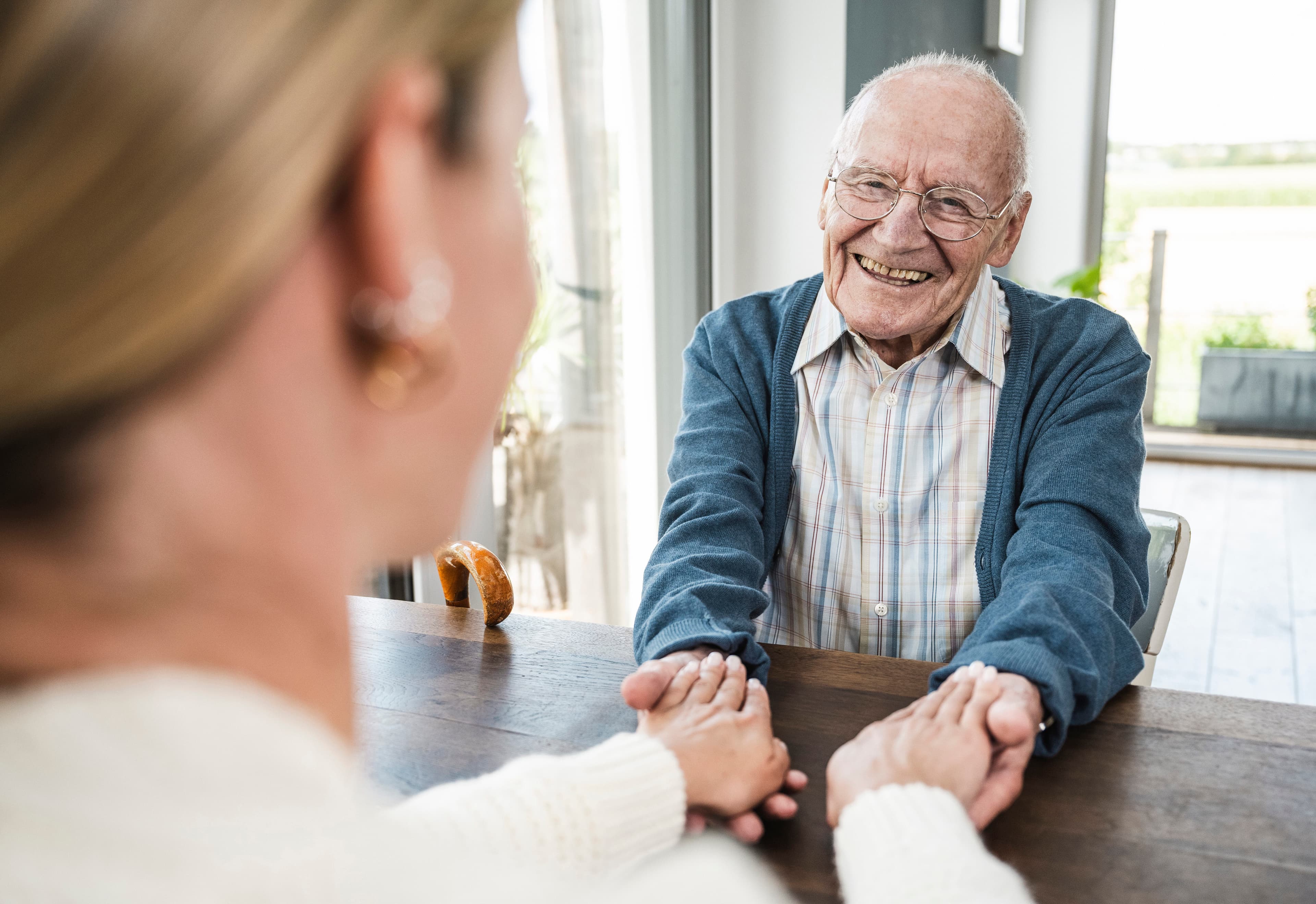 An elderly man smiles, holding hands across a table with a younger woman in a bright room. - Home Instead