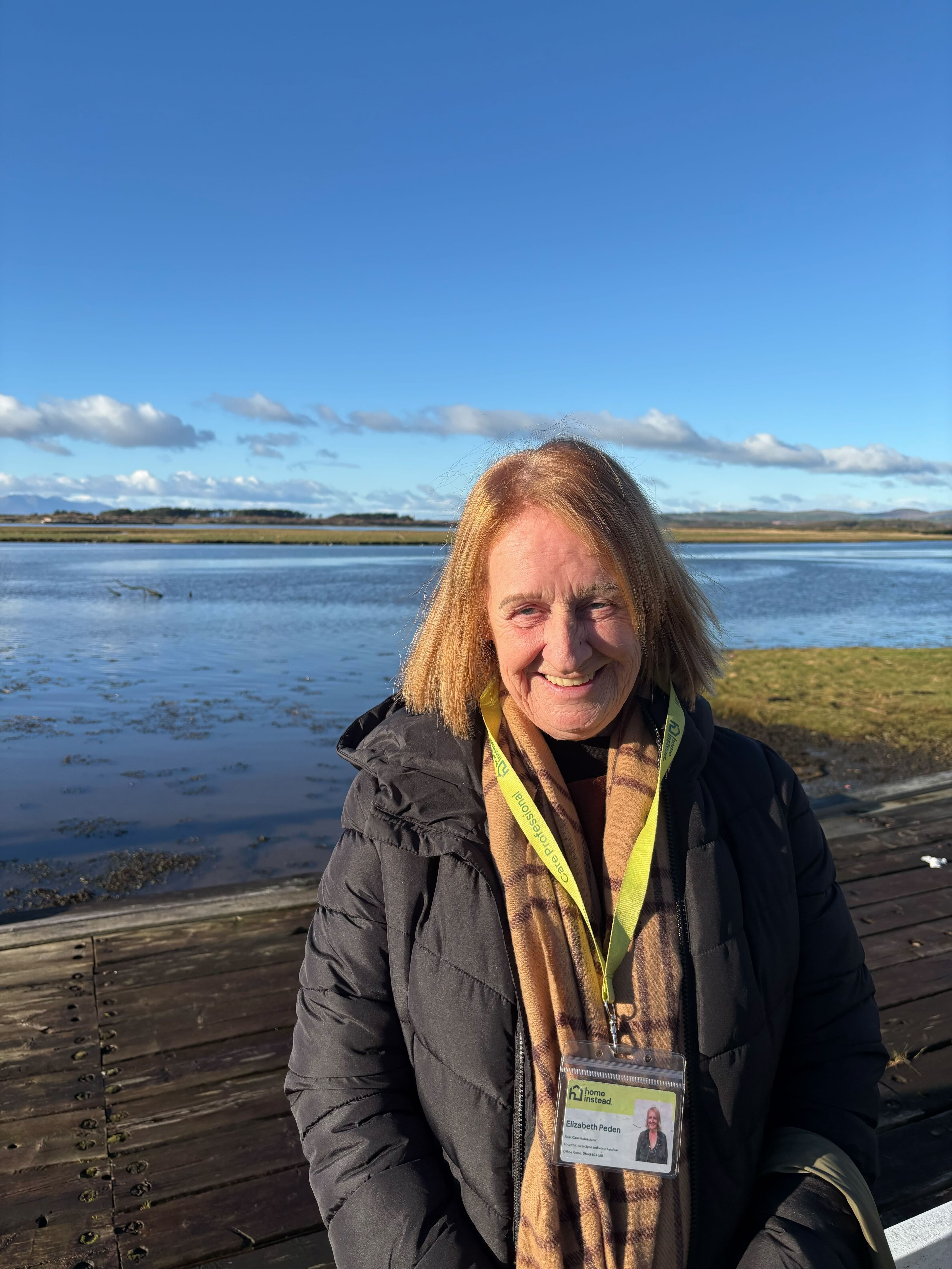 Smiling woman with a lanyard stands by a lake under a clear blue sky. - Home Instead