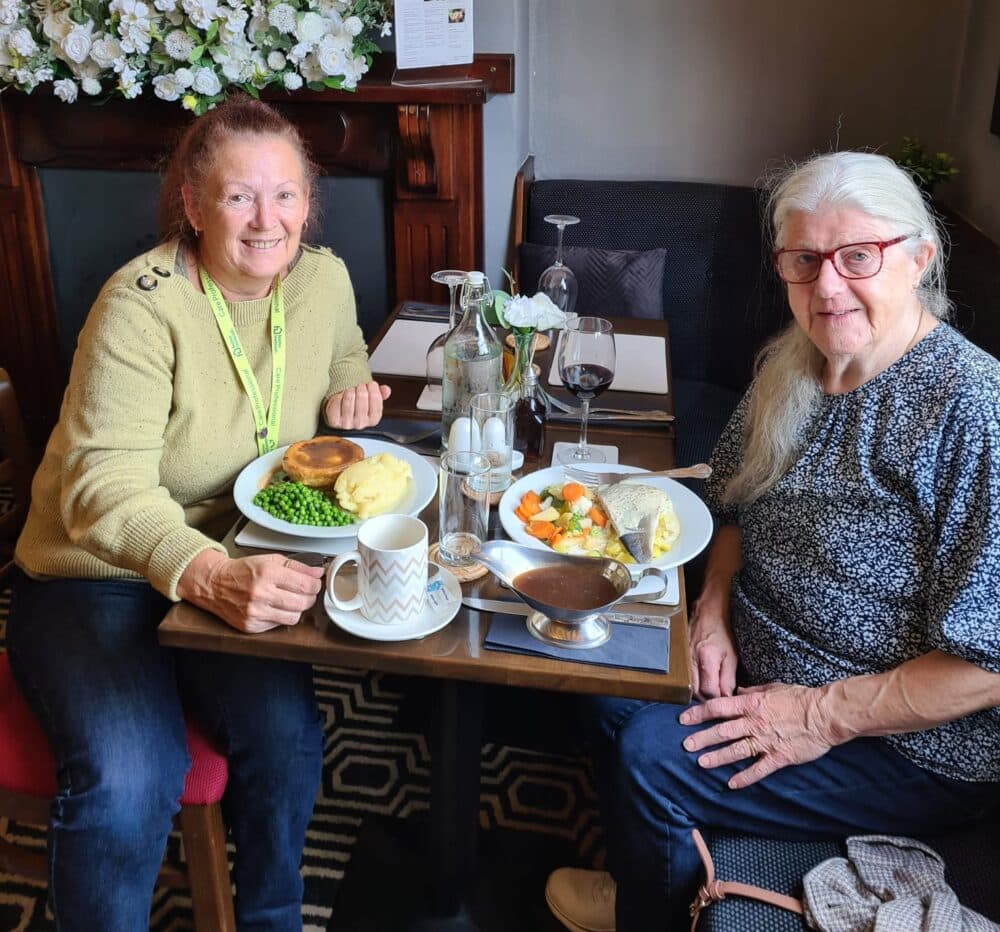 Two women smiling at a restaurant table with plates of food, drinks, and flowers in the background. - Home Instead