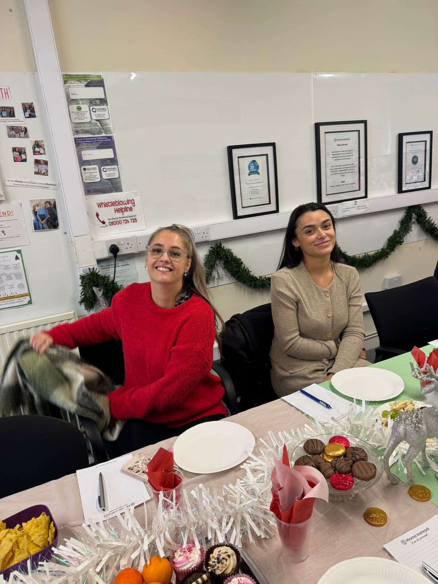 Two women sitting at a festive table with food, one smiling in a red sweater, Christmas decorations in background. - Home Instead