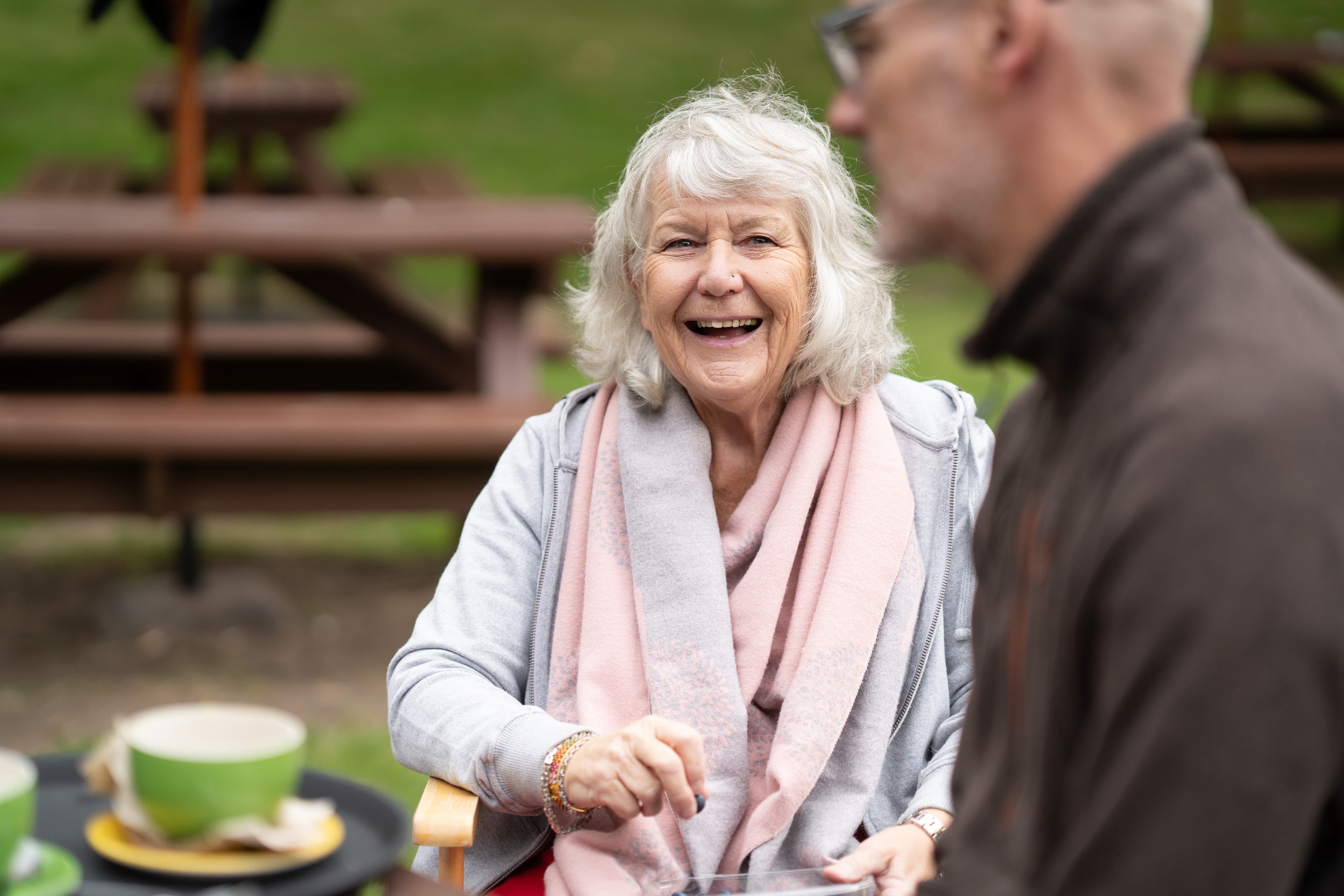 Smiling older woman sits outdoors at a picnic table, with a man in the foreground out of focus. - Home Instead